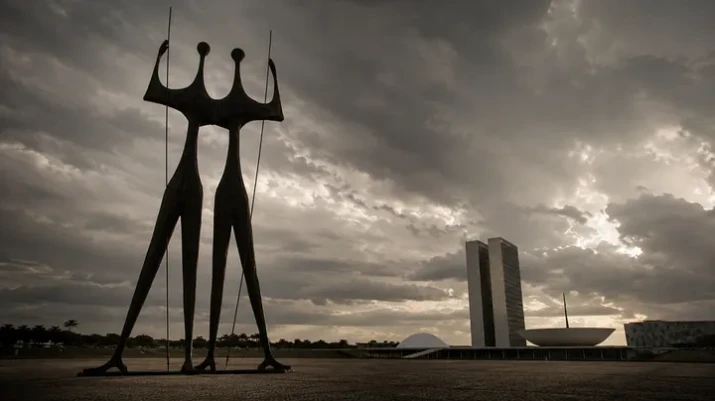 A imagem feita por IA mostra Os Candangos diante do Congresso Nacional (num angulo arquitetado pela IA), em Brasília, sob um céu pesado e dramático, criando uma atmosfera de tensão política, desencanto e desgaste institucional.